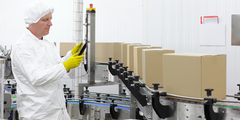 Man working on production line holding a tablet, in a white lab uniform and yellow gloves. Cardboard boxes are on the production line. 