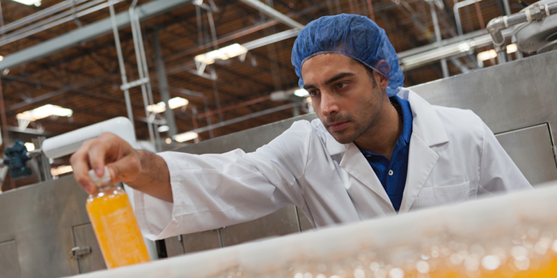 Man in white lab coat and blue hair net picking and looking at orange drink on production line